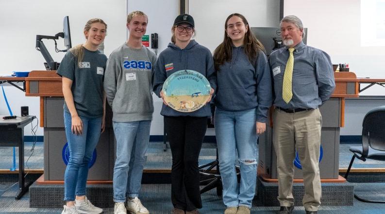 一群学生和他们的老师站在一个椭圆形的碗旁，以表彰他们在首届海洋碗比赛中获得的第一名.  