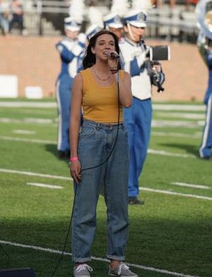 A woman sings into a microphone while standing on a football field. 
